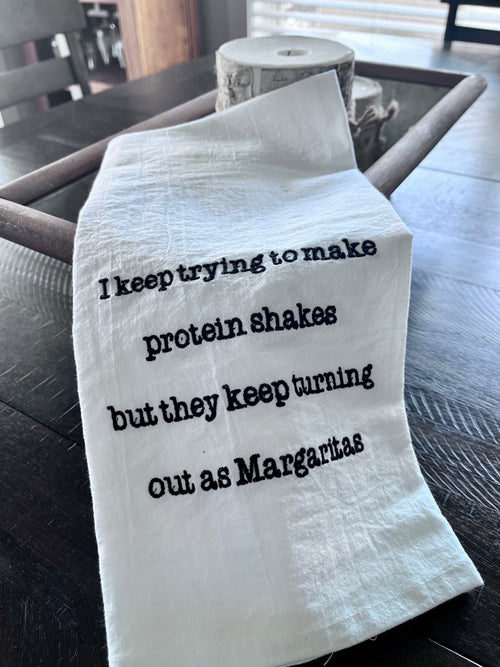A white kitchen towel with black embroidered text reading 'I keep trying to make protein shakes but they keep turning out as Margaritas', draped over a dark wooden table next to a roll of paper towels.