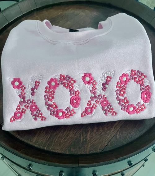 White sweatshirt with pink floral 'XOXO' design on a wooden surface