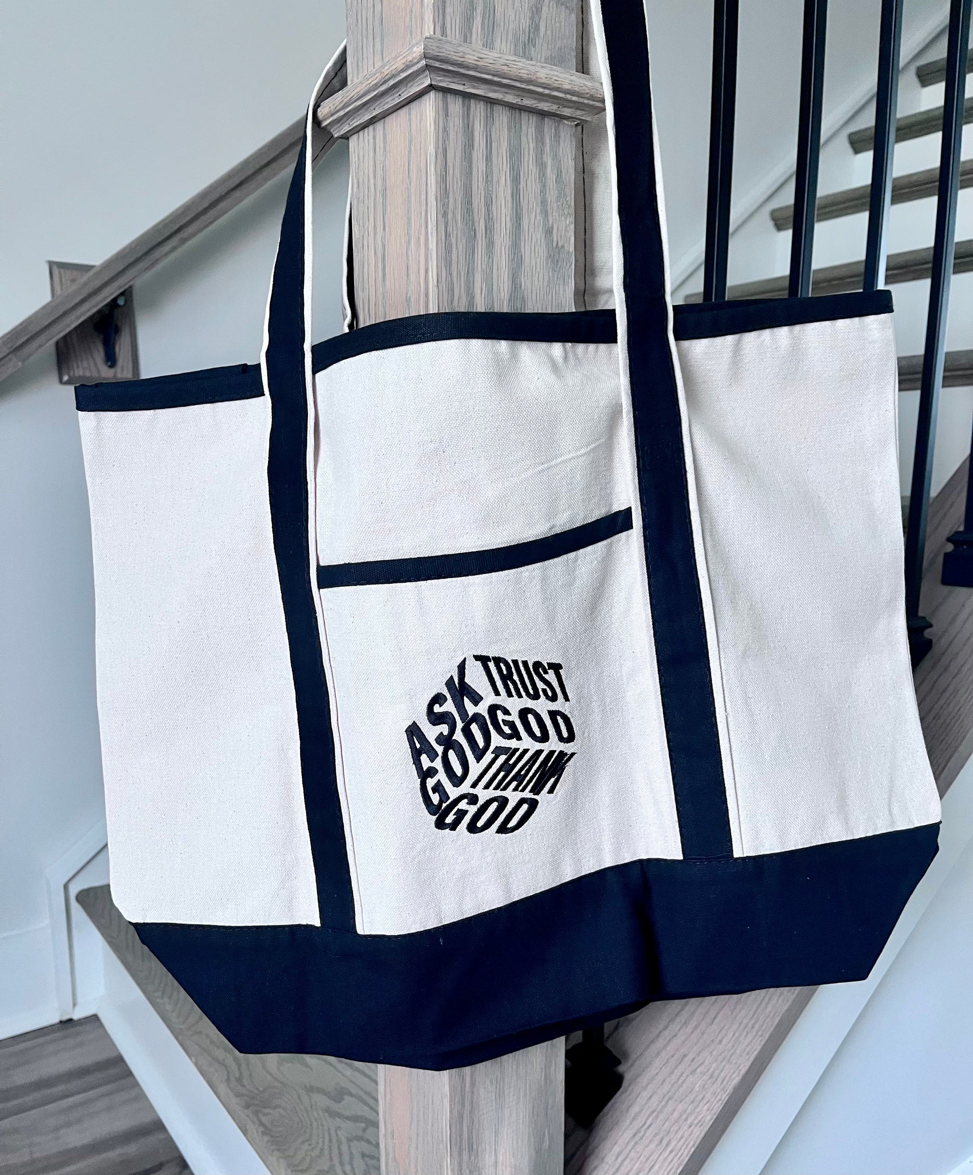 A large off-white canvas tote bag with dark navy blue trim and handles, hanging on a wooden stair railing. The front pocket features a black graphic text that reads 'ASK TRUST GOD TO SHAP ME'.