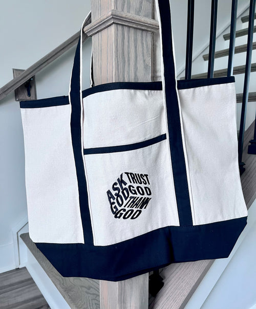 A large off-white canvas tote bag with dark navy blue trim and handles, hanging on a wooden stair railing. The front pocket features a black graphic text that reads 'ASK TRUST GOD TO SHAP ME'.