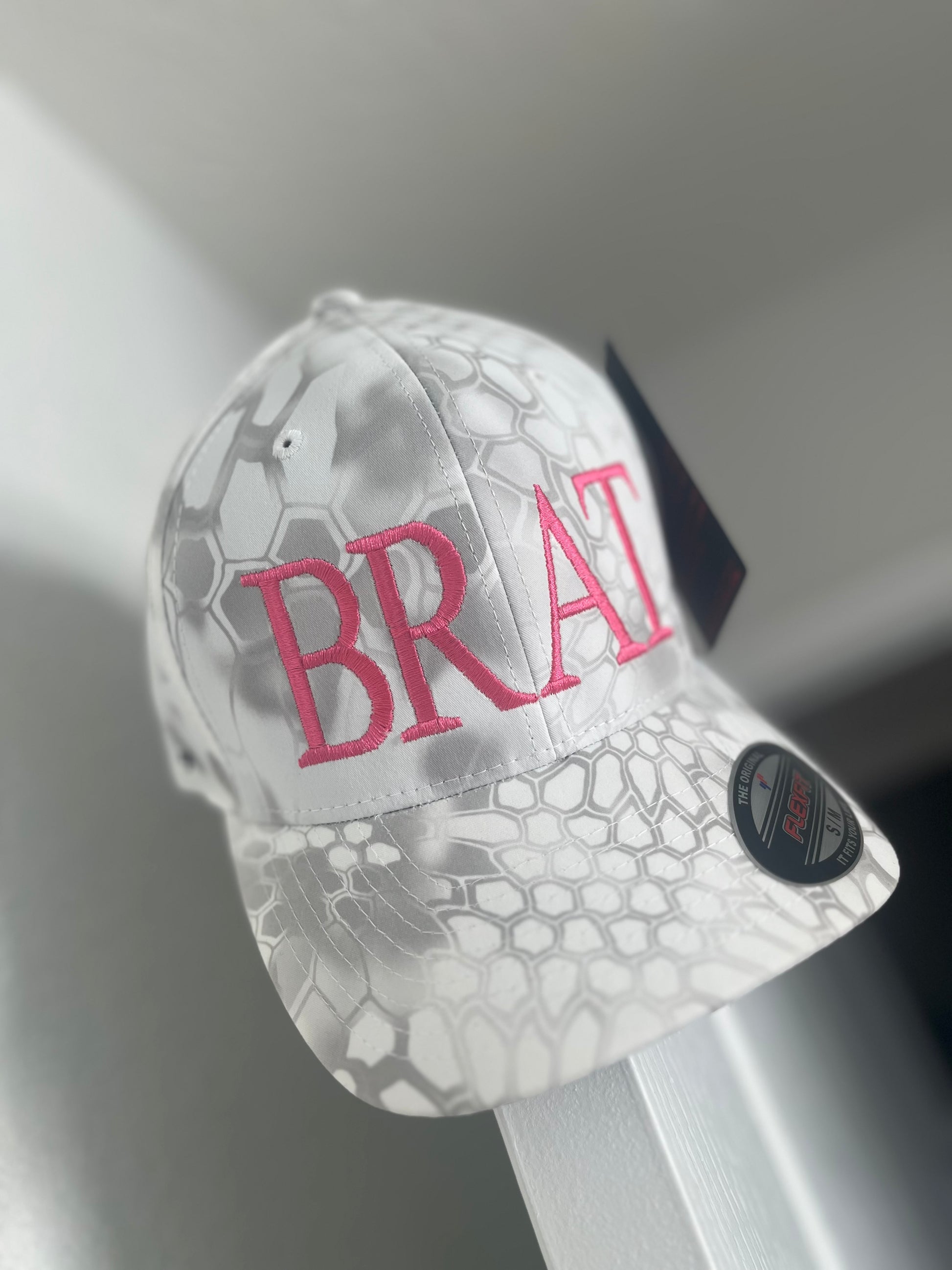 A gray camouflage Flexfit hat with the text 'BRAT' embroidered in red on the front.