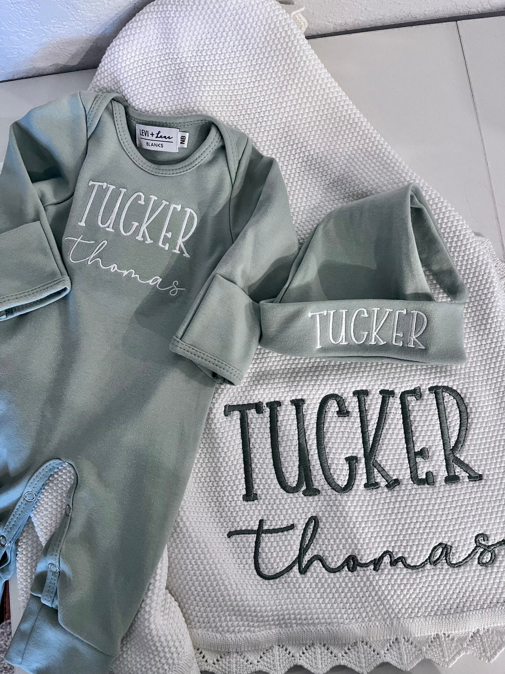 Gray baby outfit with 'Tucker Thomas' printed on it, placed on a textured surface.