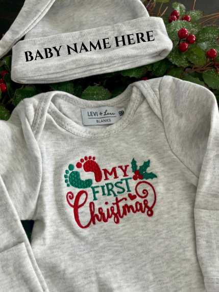 Gray baby romper with 'My First Christmas' design and matching hat on a green leaf background.