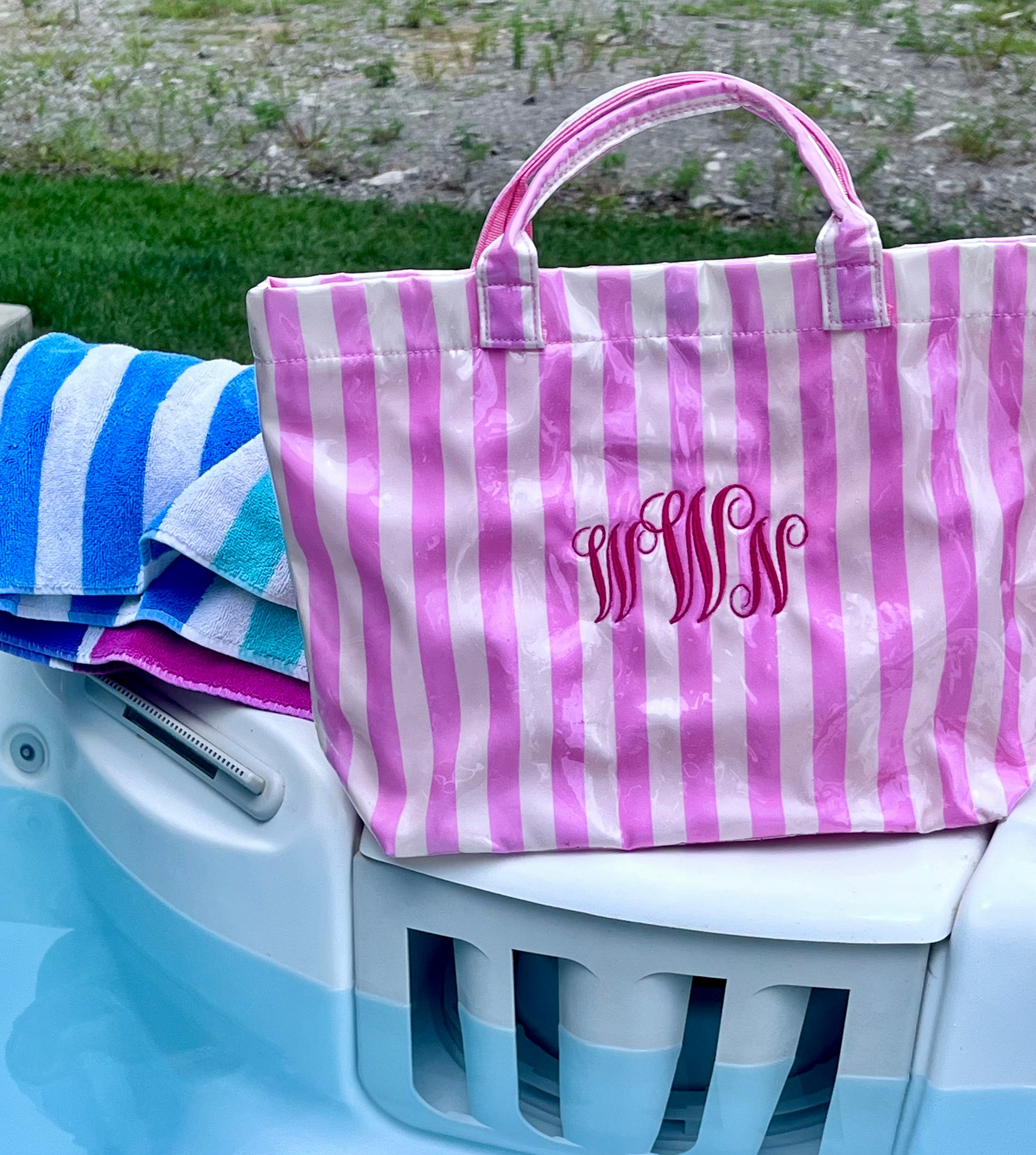 A pink and white striped pvc tote bag with a monogrammed 'MMM' logo, resting on the edge of a light blue pool next to a blue and white striped towel.