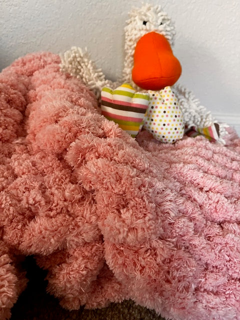 A fluffy pink chenille baby blanket draped over a surface with a stuffed toy duck sitting on top.