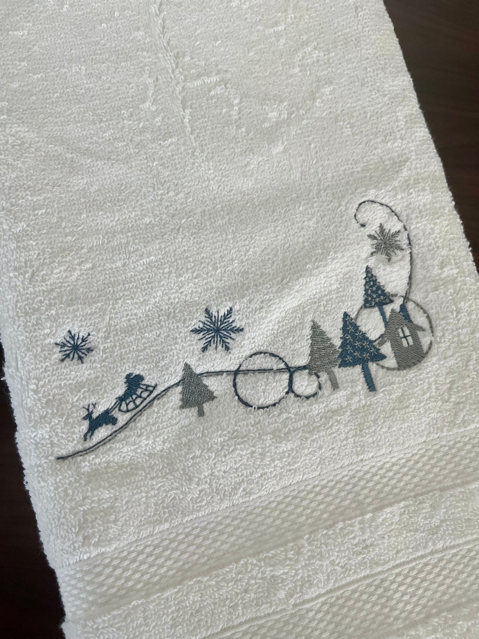 White towel with embroidered Christmas scene featuring trees, a sleigh, and a house.