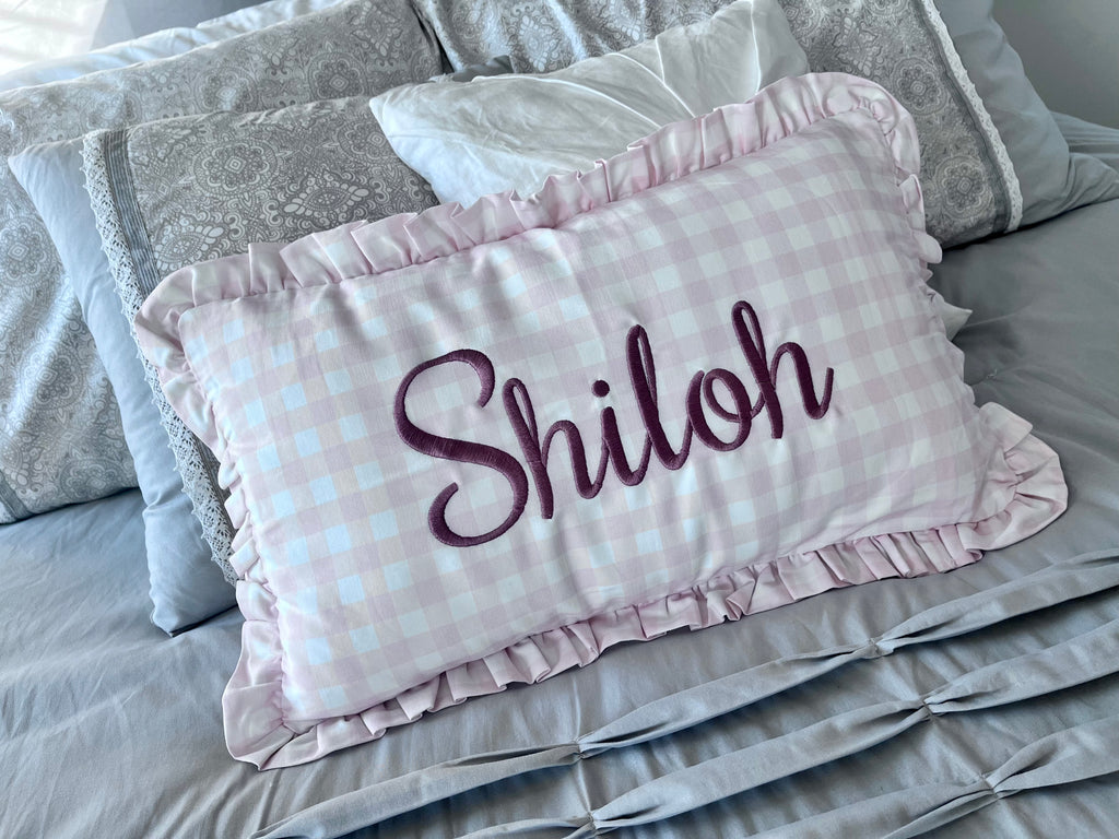 A personalized rectangular throw pillow with a light pink and white gingham check pattern, ruffled edges, and the name 'Shiloh' embroidered in dark purple script, displayed on a gray bed.