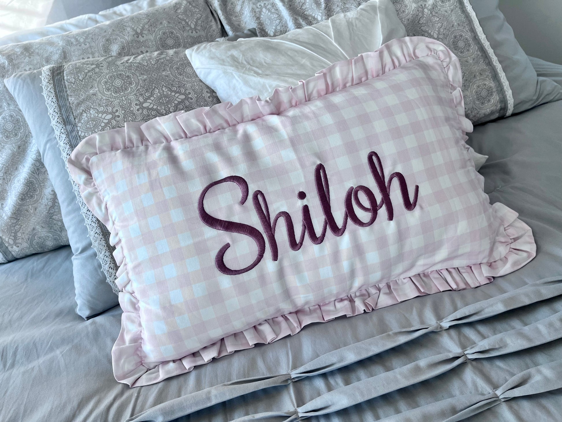 A personalized rectangular throw pillow with a light pink and white gingham check pattern, ruffled edges, and the name 'Shiloh' embroidered in dark purple script, displayed on a gray bed.
