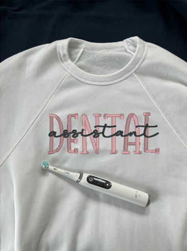 A white crewneck sweatshirt with the text 'DENTAL assistant' embroidered on the front, accompanied by an electric toothbrush.