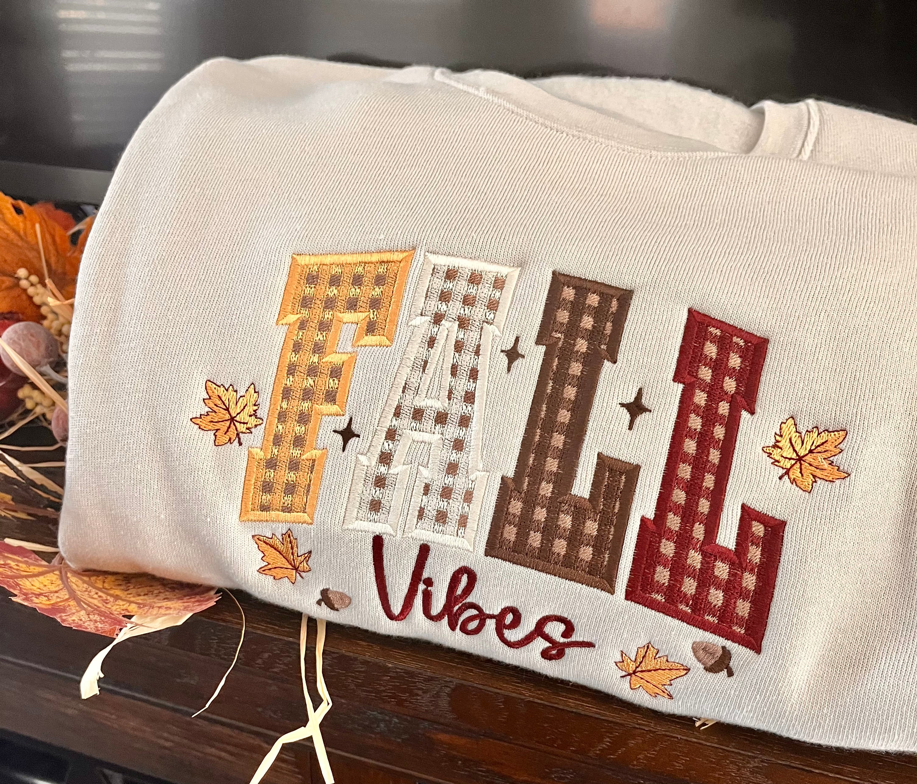 Beige pillow with embroidered 'Fall Vibes' text and fall-themed design on a wooden surface.