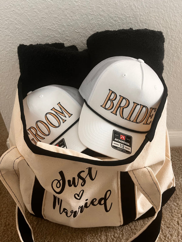 Two white caps with 'Bride' and 'Groom' text on a black towel.