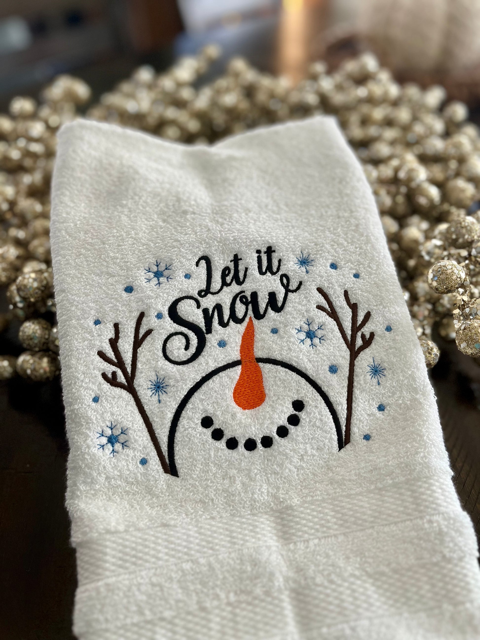 Add a touch of winter charm to your kitchen or guest bath or kitchen with this beautifully embroidered “Let It Snow” hand towel. Featuring a whimsical snowman face with twig arms, a carrot nose, and scattered snowflakes, this towel brings cozy holiday cheer to any space.