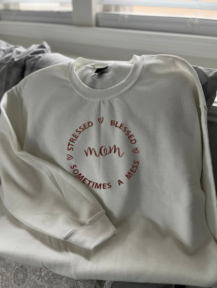 White crewneck sweatshirt with the text 'MOM Stressed Blessed Always A Mess' embroidered in a circular pattern with mauve thread.