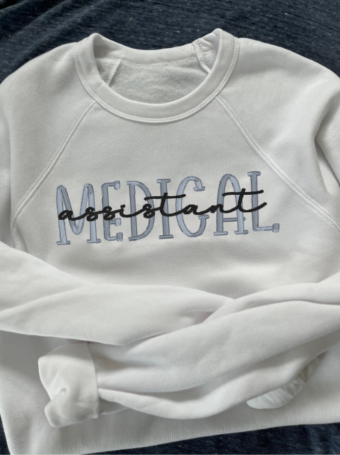 White crewneck sweatshirt with 'MEDICAL assistant' embroidered on the front in two different colors of thread.