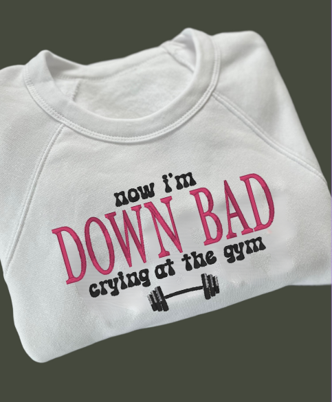 White embroidered crewneck sweatshirt with the text 'now I'm DOWN BAD crying at the gym' printed on the front.