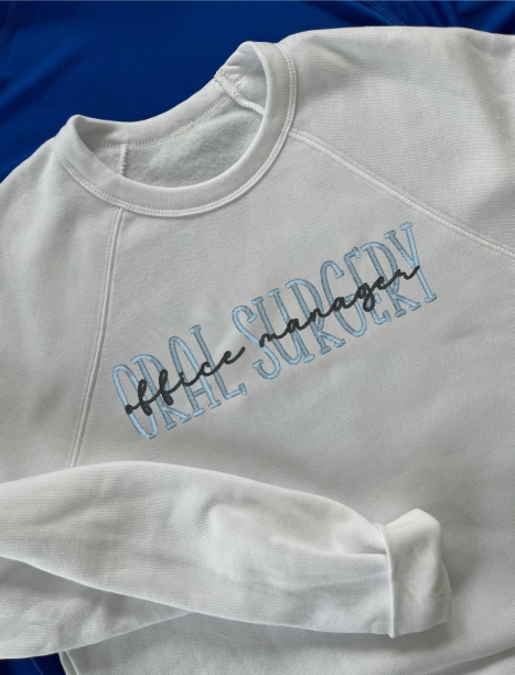 A white embroidered crewneck sweatshirt with the text 'ORAL SURGERY' and additional script embroidered on the front.