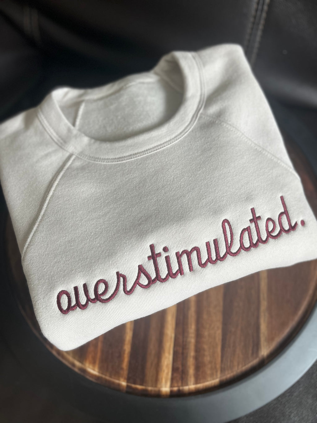 A beige crewneck sweatshirt with the phrase 'overstimulated.' embroidered in red on the front.