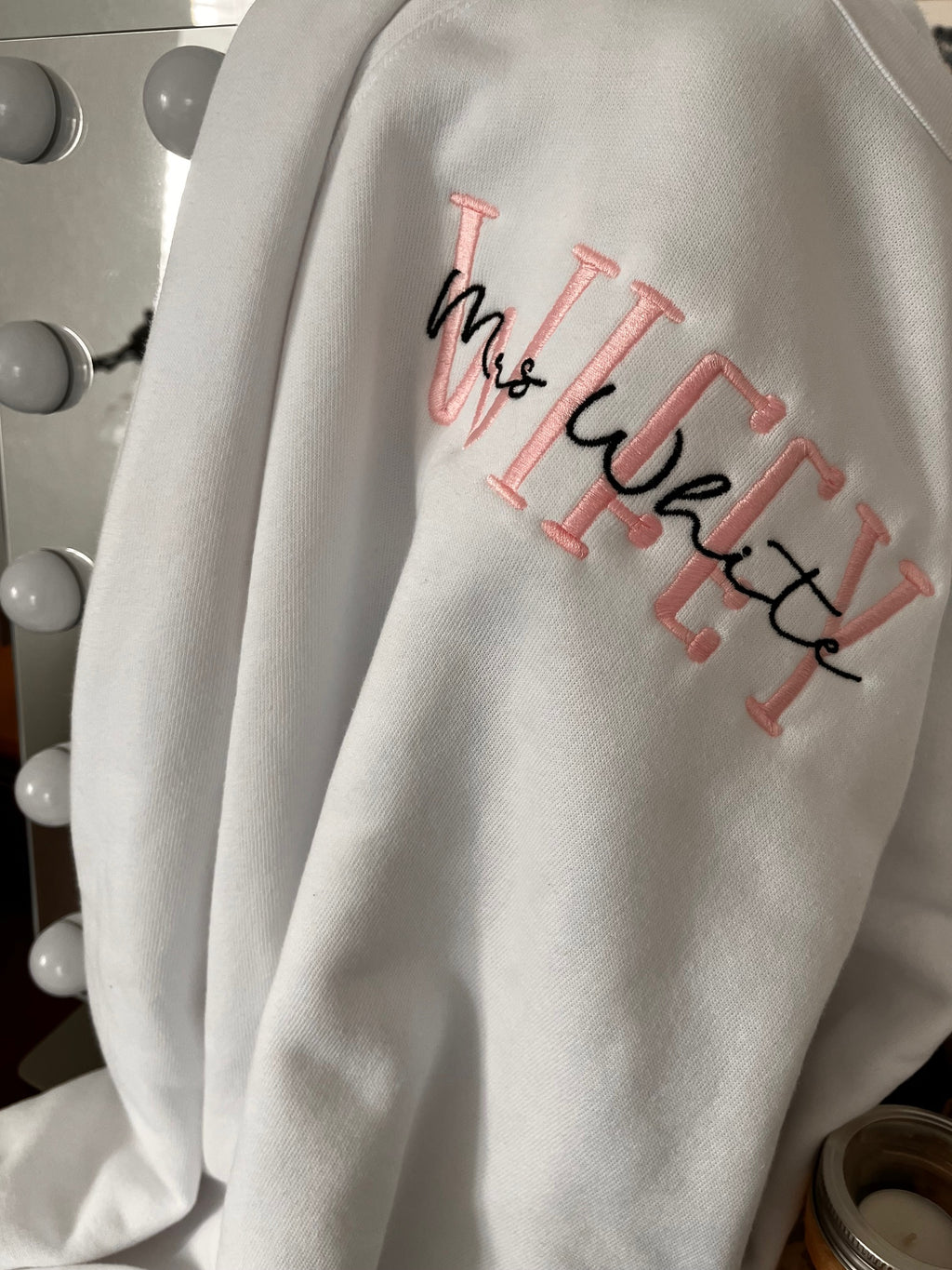 A gray crewneck sweatshirt with the word 'WIFEY' embroidered in pink and black on the sleeve.