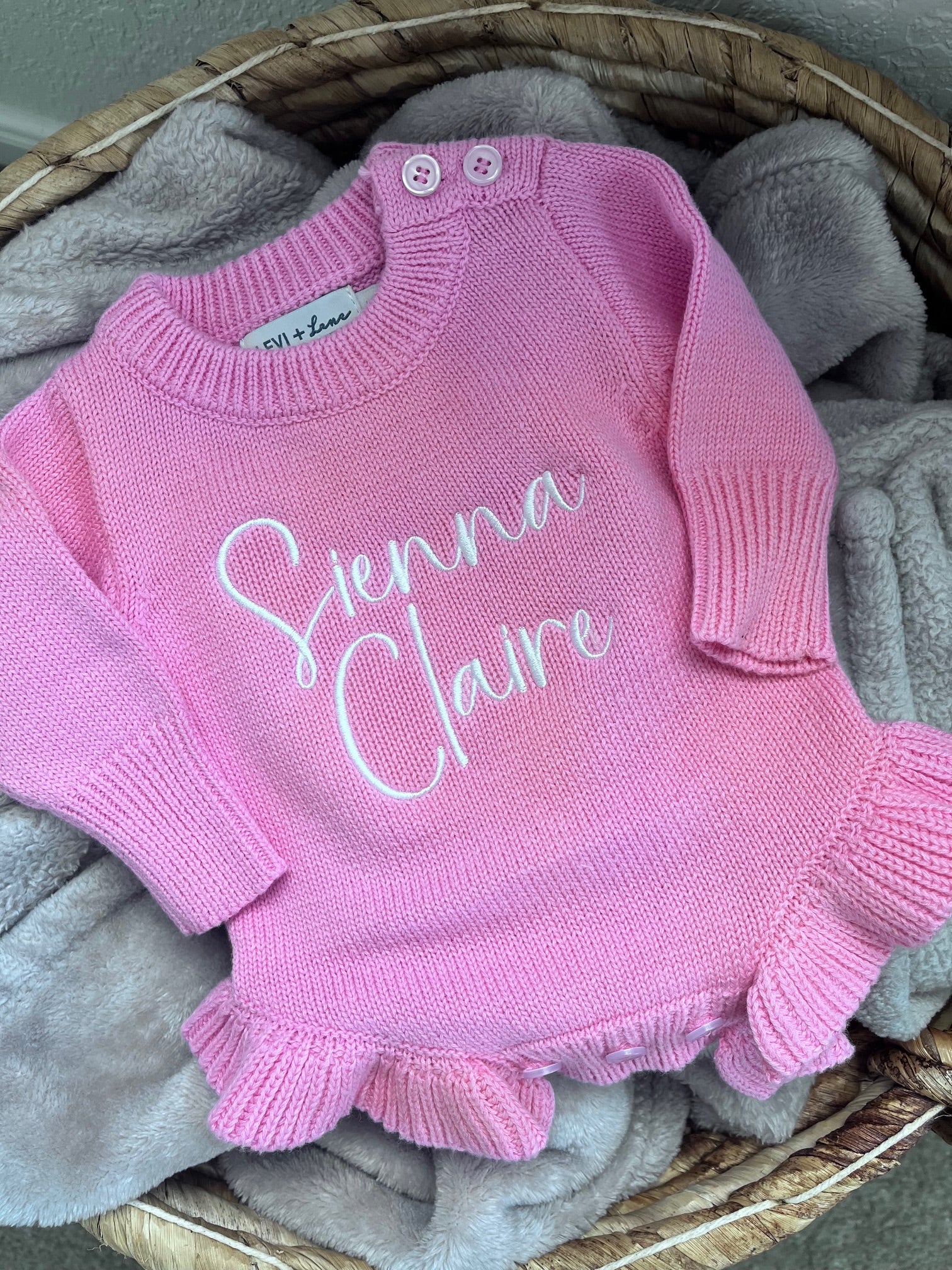 A pink knit baby romper with long sleeves and ruffled hems, featuring the personalized name 'Stenna Claire' embroidered in white script across the chest. The romper is displayed in a wicker basket lined with a soft gray blanket.
