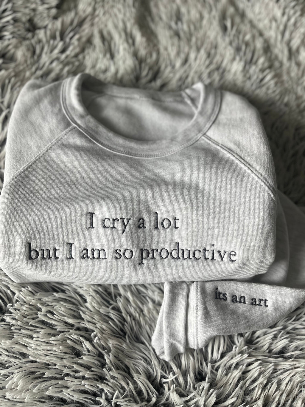 A gray crewneck sweatshirt with the text 'I cry a lot but I am so productive' written on the front and 'in an art' on the sleeve, displayed on a plush surface.