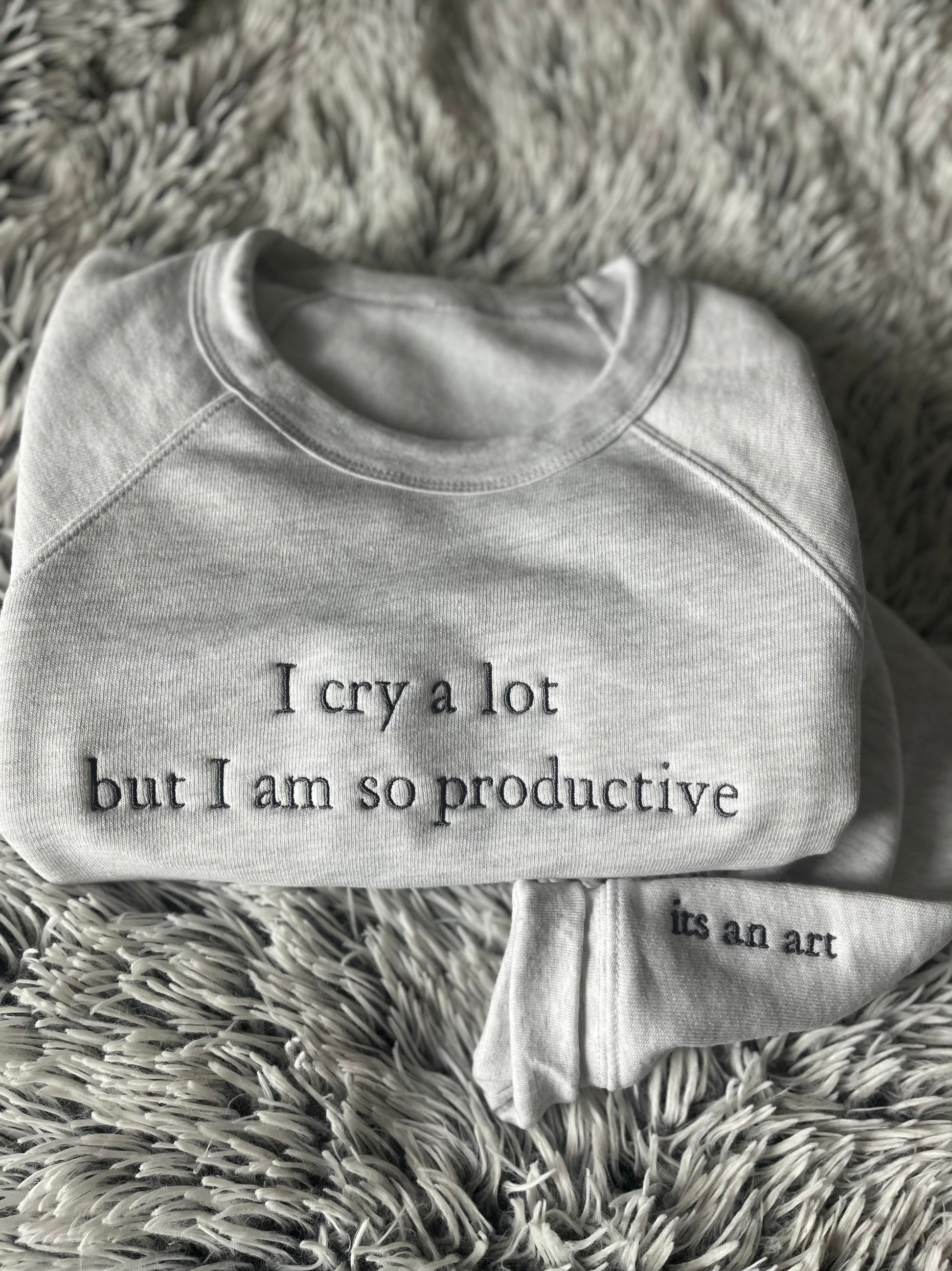 A gray crewneck sweatshirt with the text 'I cry a lot but I am so productive' written on the front and 'in an art' on the sleeve, displayed on a plush surface.