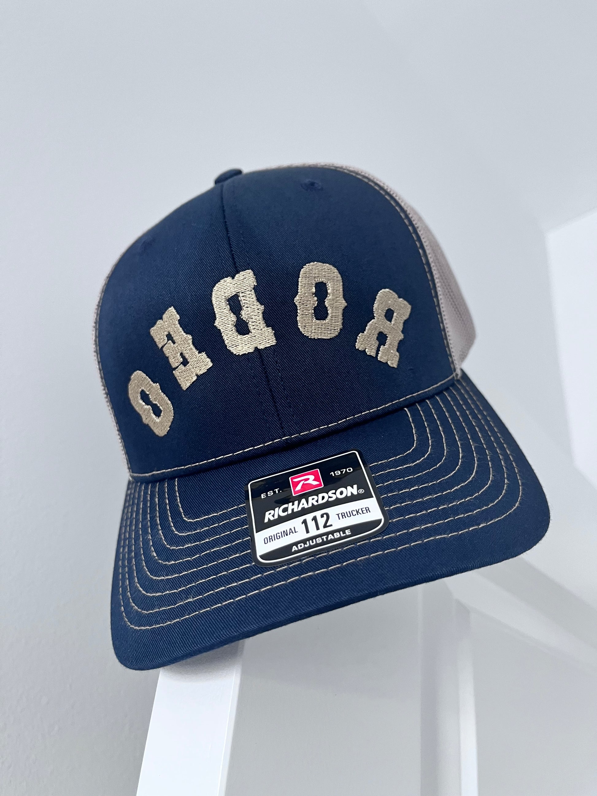 A Richardson 112 trucker cap in navy blue with beige embroidered lettering on the front and a matching beige mesh back, displayed on a white stand.