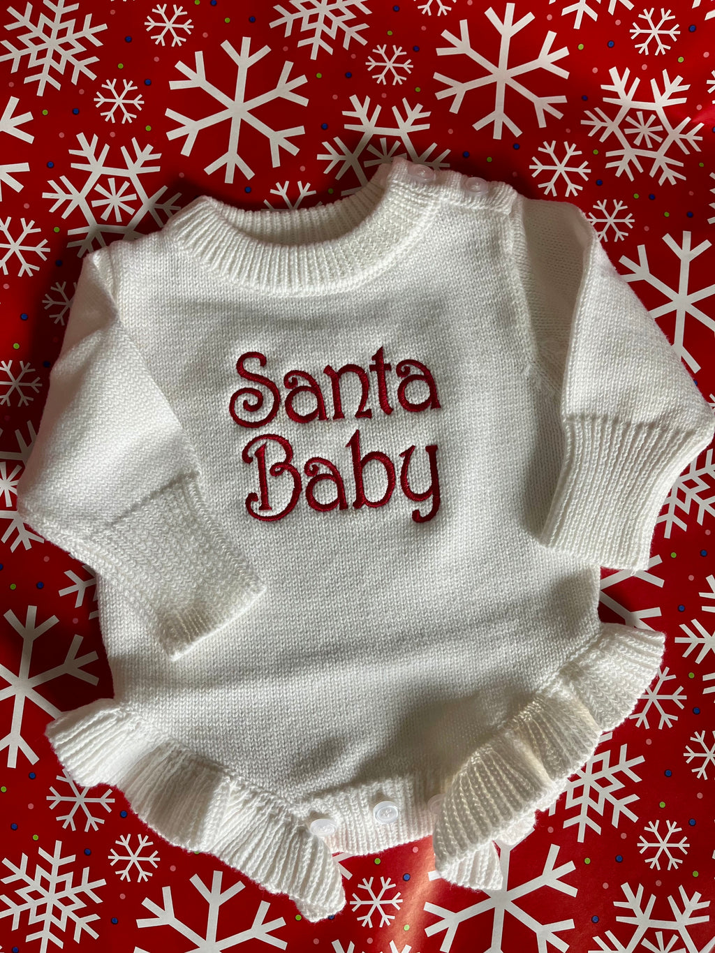 White baby sweater with 'Santa Baby' text on a red background with snowflakes