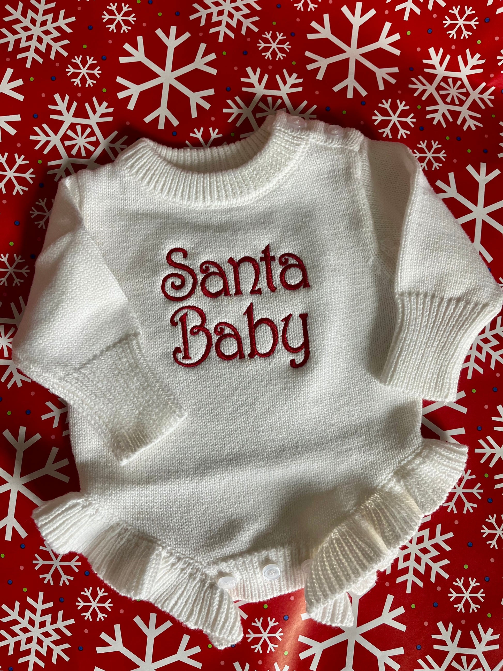 White baby sweater with 'Santa Baby' text on a red background with snowflakes