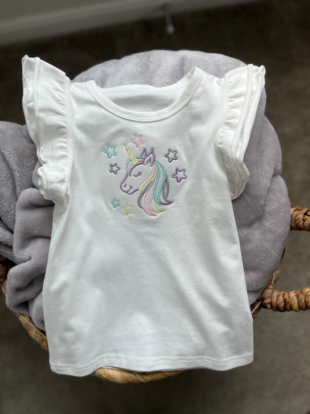 A white t-shirt with ruffled sleeves featuring a unicorn and stars embroidery, displayed on a grey background.