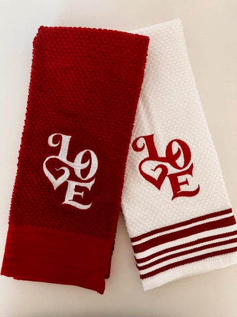 Two kitchen towels, one red and one white, each embroidered with the word 'LOVE' in a heart shape.