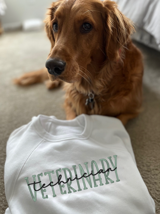 A white crewneck sweatshirt with 'VETERINARY' and 'technician' text embroidered on the front, accompanied by a golden retriever lying down in the background.